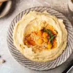 Cremiger veganer Hummus in einer Keramikschale, garniert mit ganzen Kichererbsen, weißem Sesam, aromatischem Öl und einem frischen Kräuterzweig by NAR Kitchen
