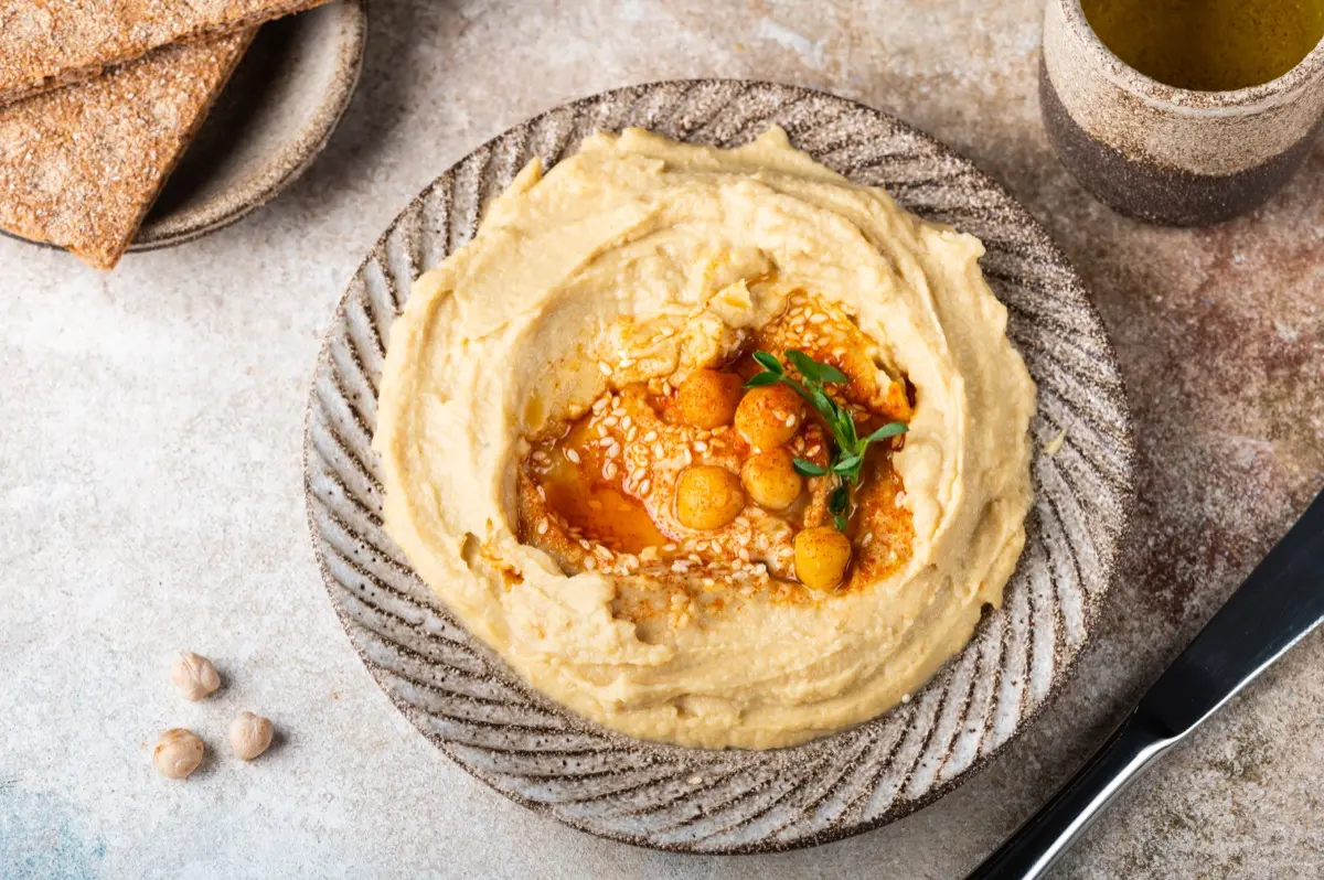 Cremiger veganer Hummus in einer Keramikschale, garniert mit ganzen Kichererbsen, weißem Sesam, aromatischem Öl und einem frischen Kräuterzweig by NAR Kitchen